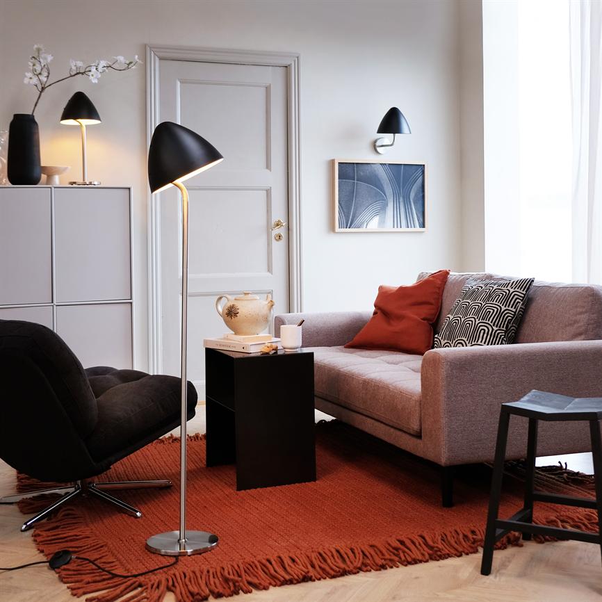 Floor lamp illuminates a small black side table with a jug and a mug. Context: light gray sofa with patterned and red cushions, black armchair and red fringed carpet in bright living area.