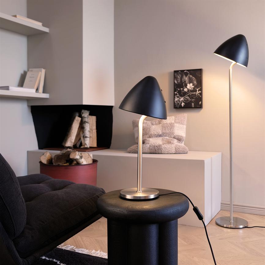 Black table lamp with curved shade stands and shines softly on a round black side table, in front of a dark sofa; behind it is a matching floor lamp, white sideboard with rug and firewood basket.
