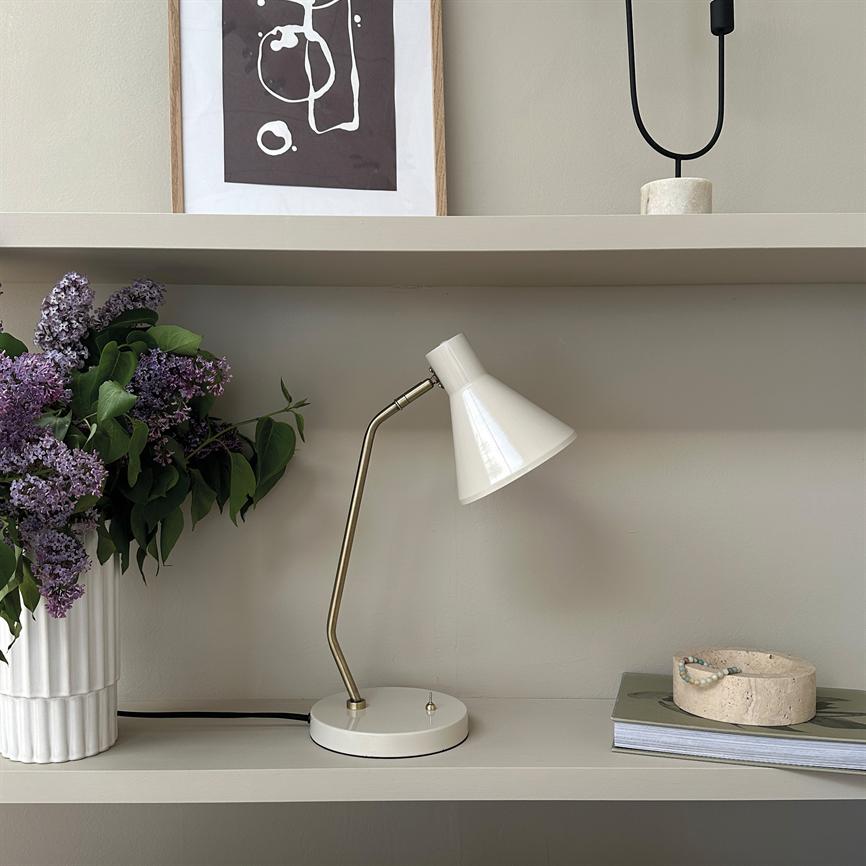 A white table lamp points obliquely downwards and is switched off on a shelf; on the left a vase with purple lilacs, above the shelf a framed abstract print, on the right books and bowl.