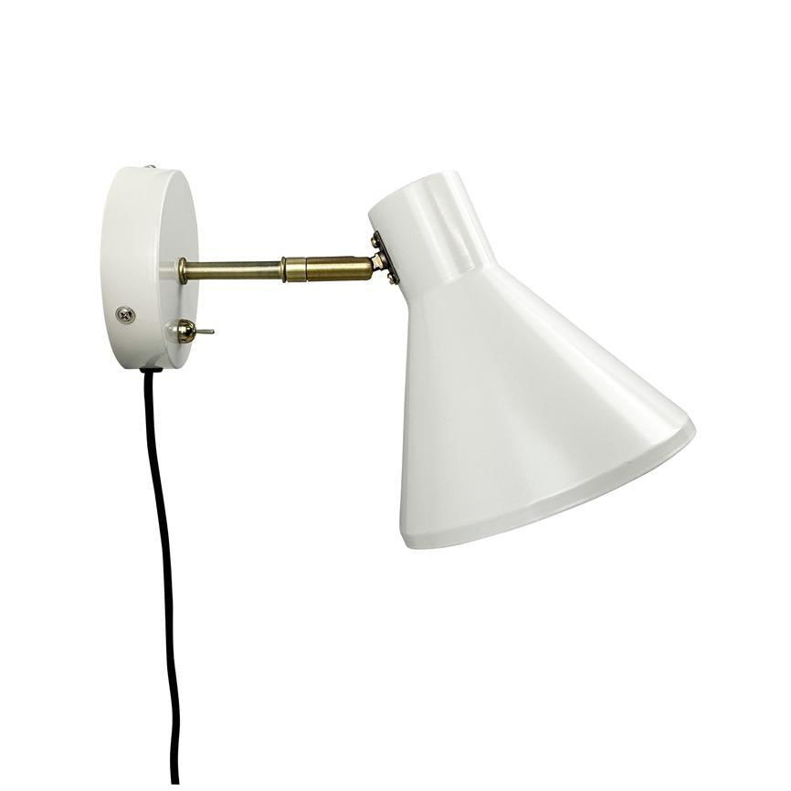 Wall lamp mounted on a round white wall plate with a short brass arm; white conical screen is angled downward and a black cord hangs freely against a neutral white background.