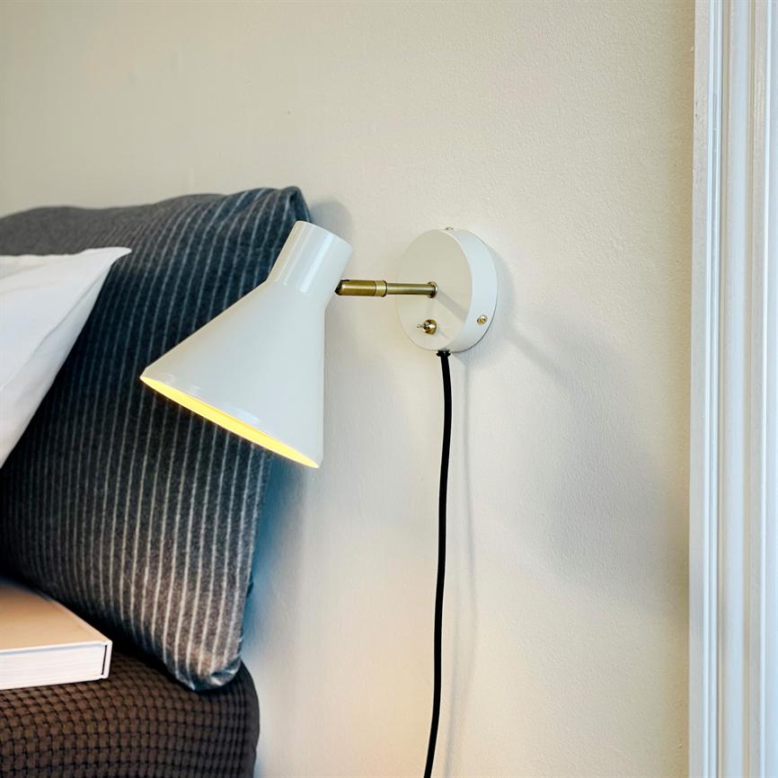 White wall lamp lights up and casts warm light down towards the headboard; mounted on light wall with black cord, by a bed with dark grey, striped pillow and a book on the edge of the bed.