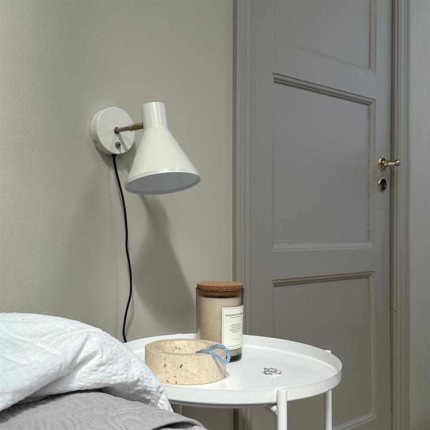 White wall lamp hangs unlit above a round bedside table with a glass scented candle, cork lid, a stone tray and jewellery; quiet bedroom and closed door. No clear readable text.