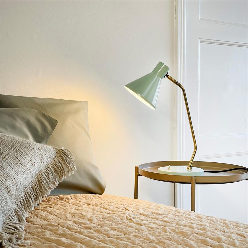 Green metal table lamp shines above a round brass bedside table next to a bed with a beige quilted cover and knitted pillow in front of a white wall.