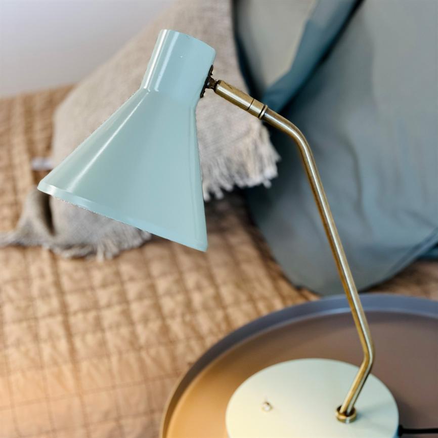Light blue table lamp bends its brass arm and directs the shade downwards over a round bedside table; in the background you can see a quilted bedspread, a gray pillow and a braided rug.