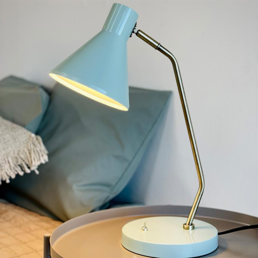 A blue metal table lamp shines downward from a curved brass arm; it stands on a round bedside table next to a bed with grey-blue pillows and a frayed white throw blanket.