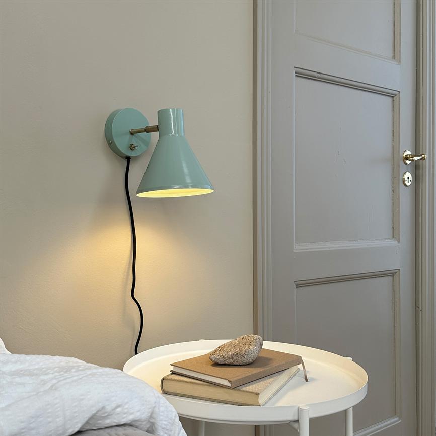 A mint green wall lamp shines softly over a round white side table with two books and a stone, next to a bed and a light gray paneled door on a beige wall.