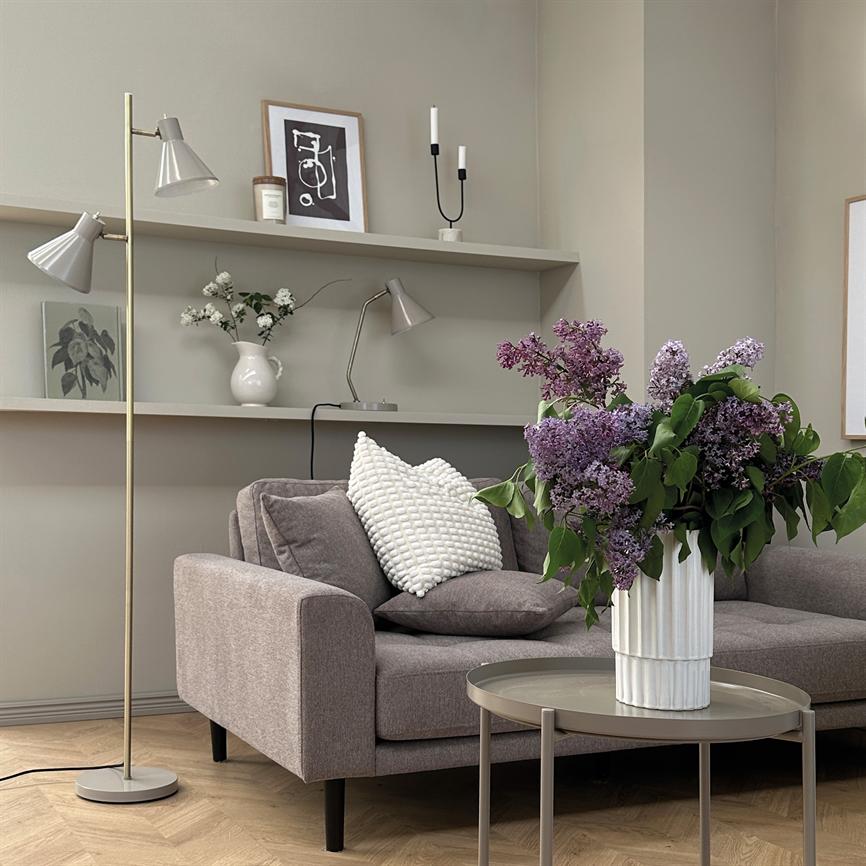 A bouquet of purple lilacs stands in a white vase on a round coffee table; in the background is a gray sofa with cushions, shelves and a floor lamp in a quiet living room.