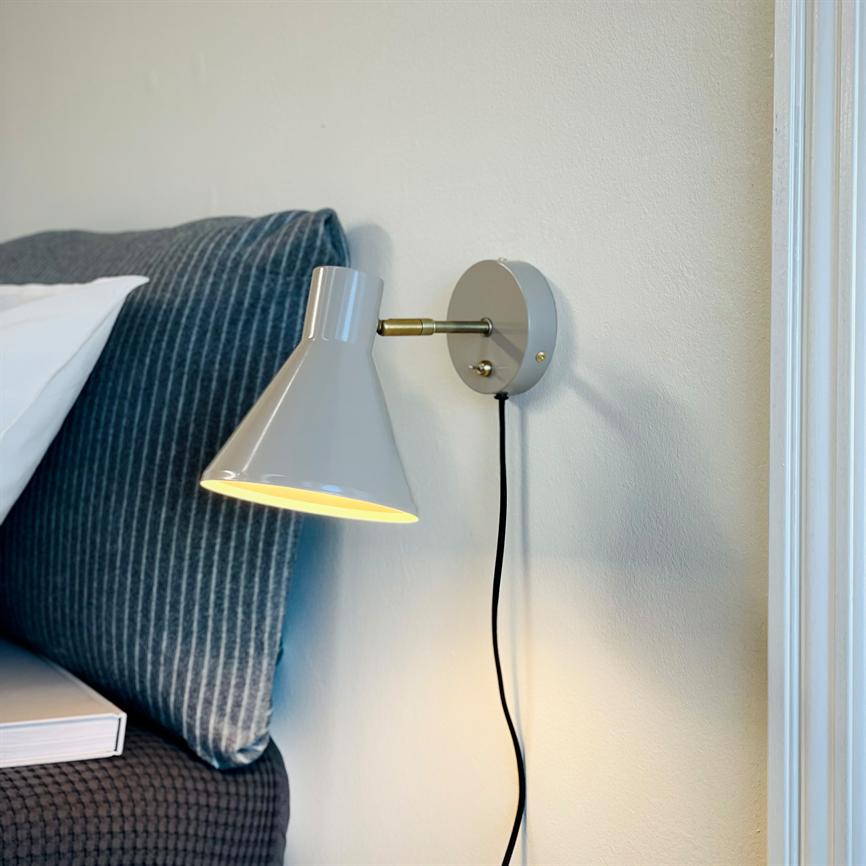 Wall lamp: a grey, conical lamp lights downwards; mounted by a striped blue pillow and the headboard of the bed, with black cord hanging along a light wall.