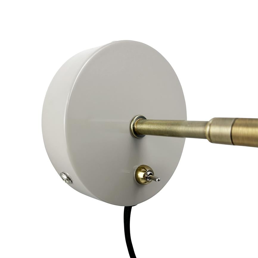 Round white wall plate with a brass rod piercing through the center; a small brass rocker switch sits at the bottom, black wire hangs down in a neutral white background.