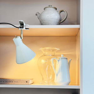 A white coffee pot is lit by a clamp lamp; it stands next to a clear glass jar; on the shelf above a gray ceramic jug and below a book. Text on spine: "THE KINFOLK TABLE".