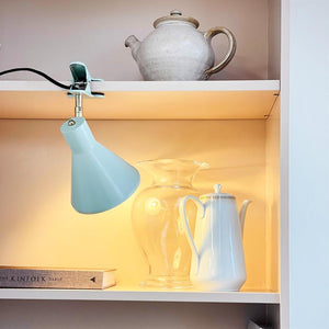 A mint green clamp lamp shines on a clear glass jar and a white jug on a lighted shelf, below a ceramic teapot; book spine with the text THE KINFOLK TABLE.
