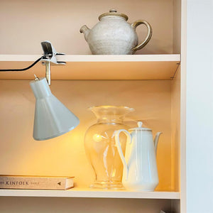 White porcelain coffee pot stands next to a clear glass vase, lit by a gray clamp lamp, on a light beige built-in shelf with a clay pot on top. 
 Text: THE KINFOLK TABLE