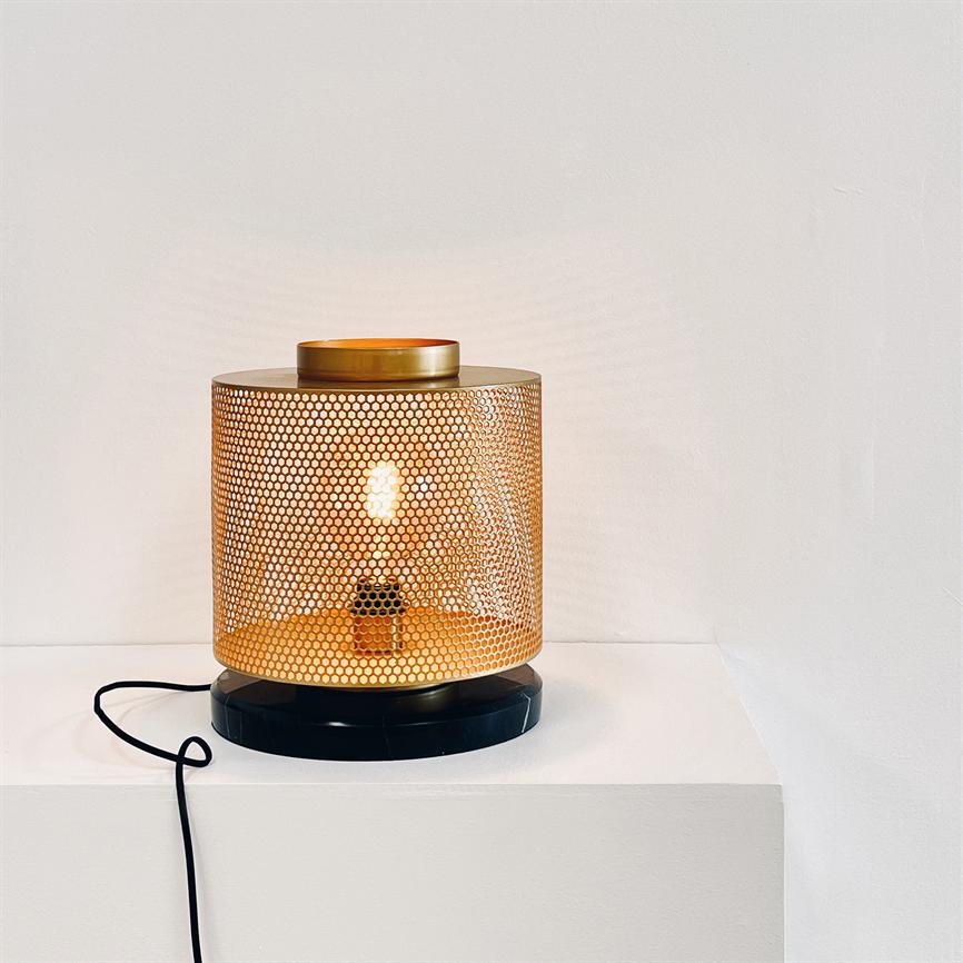 A cylindrical gold-colored table lamp emits warm, patterned light through a perforated metal shade; it stands on a white plinth in front of a light wall, with a black cord at the base.
