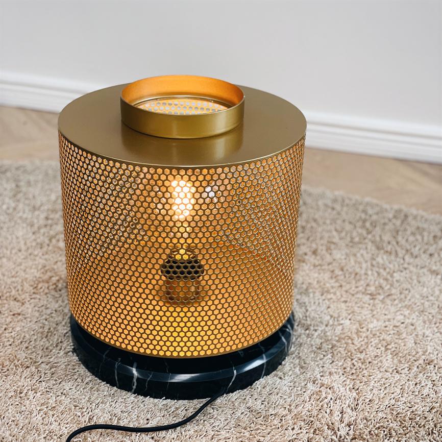 Cylindrical gold colored table lamp with perforated hexagon pattern, emitting warm glow from a visible bulb, stands on black marble base placed on light carpet in front of a white wall.