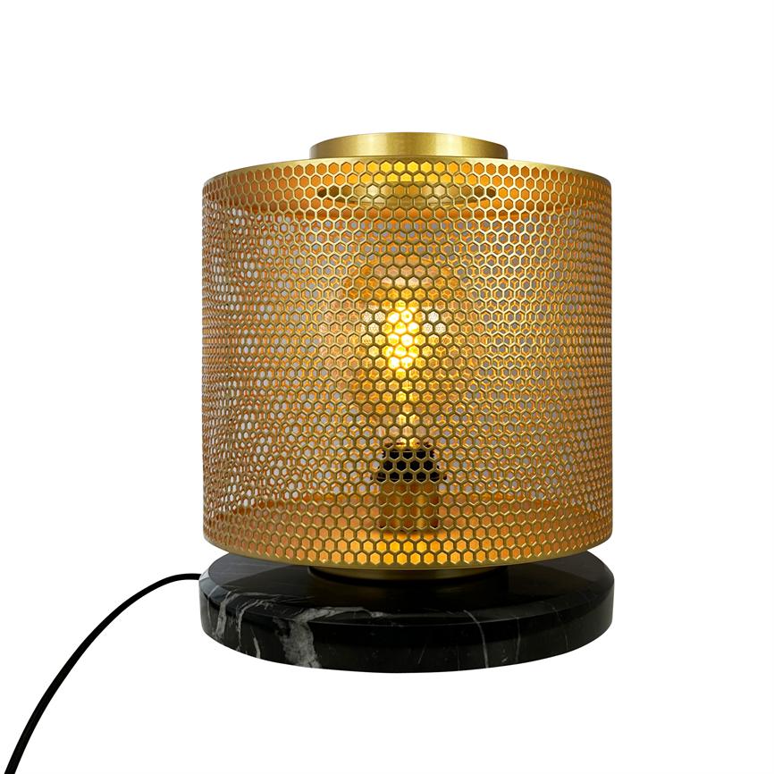 Gold-lit table lamp with perforated, gingerbread-like cylinder shade, where a lighted bulb shines through the pattern; stands on round black marble base with visible black cord on white background.