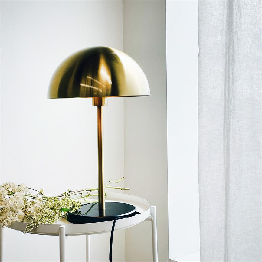A gold-colored dome-shaped table lamp stands on a round white side table; black base and cord, white flowers lie beside, by a light window with a white curtain.