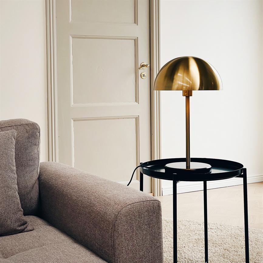 A brass dome-shaped table lamp stands on a round black side table next to a gray sofa in a bright, minimalist living room with a sliding door, light wooden floor and light carpet.