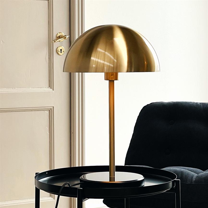 A round brass lamp stands on a black round side table; its domed screen rests on a slender brass rod. The background is a beige door and a dark, upholstered armchair.