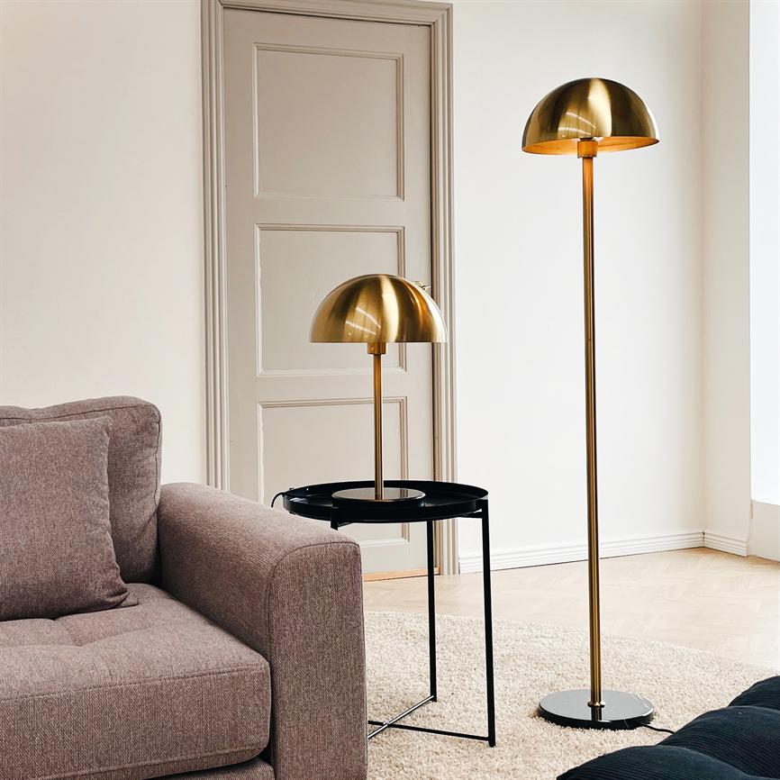 Two gold lamps stand by a gray couch; one on a black side table, the other as a floor lamp on the wooden floor, surrounded by light walls, a beige door and a bright carpet.