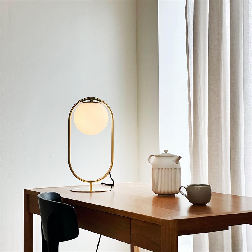 En oval bordlampe med rund, oplyst glaskugle står på et træbord og lyser; ved siden står en ribbet keramikkande og en kop, foran en sort stol ved et vindue med lyse gardiner.