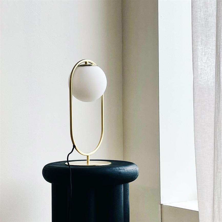 En moderne bordlampe med hvid glaskugle i en forgyldt oval ramme står slukket på en sort rund skammel ved et lyst vindueshjørne med beige vægge og hvidt gardin.