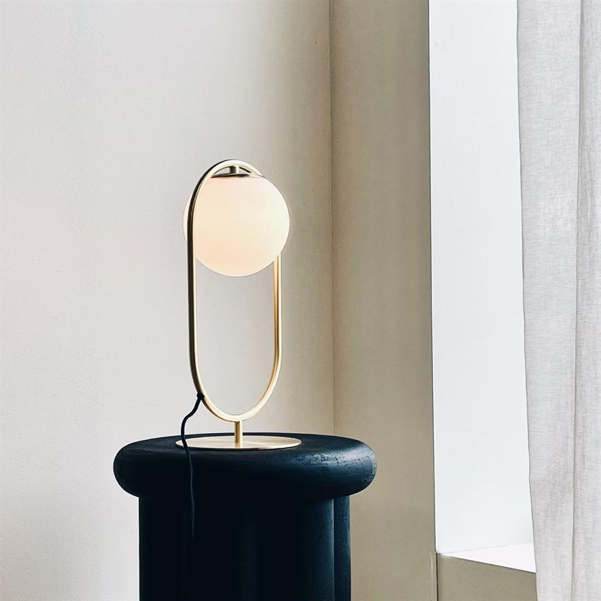 En moderne bordlampe med gyldent ovalt stativ og opal glaskugle udstråler blødt, varmt lys, stående på en rund, mørk skammel i et lyst hjørne ved et gardinbeklædt vindue.
