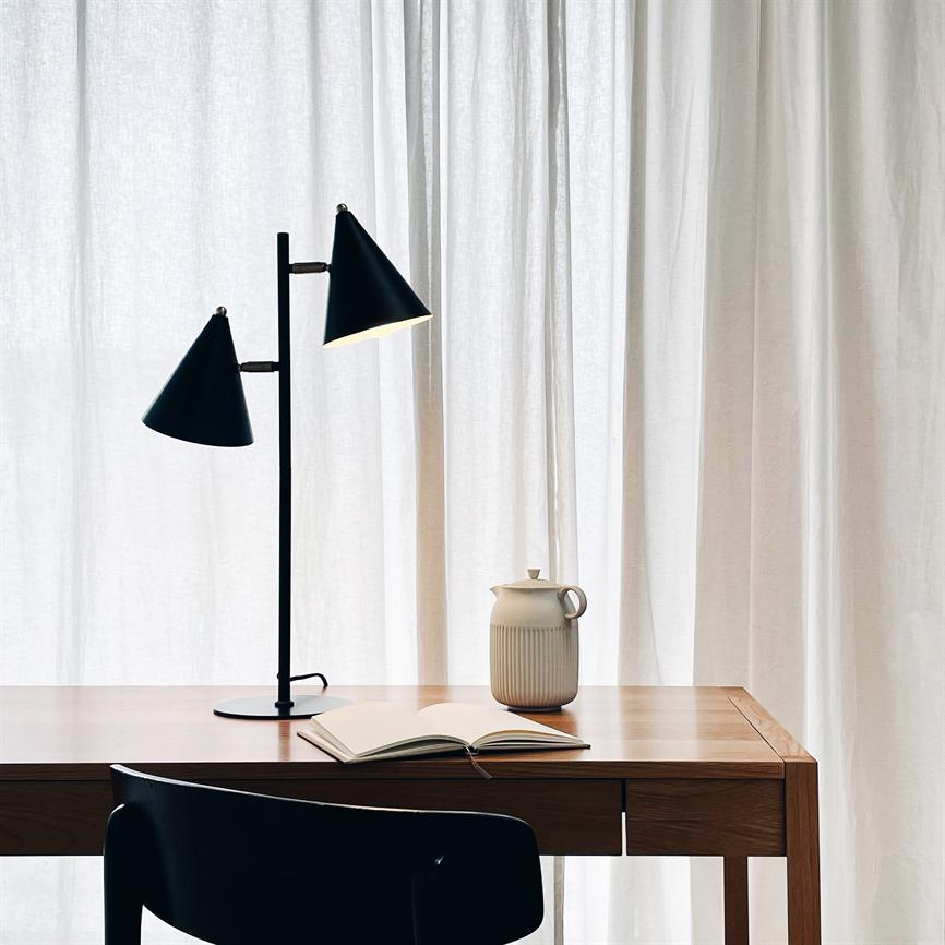 A black double cone lamp points down towards the table; on the wooden table is an open notebook and a ceramic jug, a black chair in front and soft white curtains in the background.