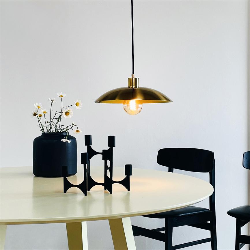 A golden hanging lamp shines above a round, bright table; a black vase with daisies and a black geometric candlestick stand on the table in a minimalist dining room with black chairs.