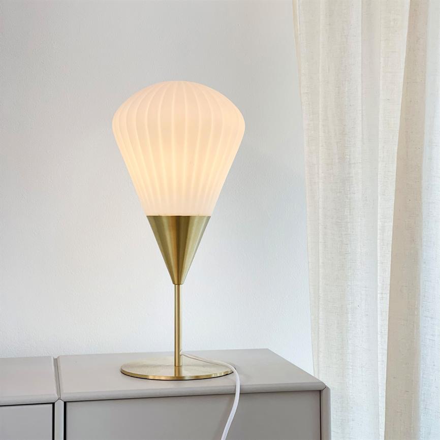 Bordlampe med opalhvid, ribbet skærm og messingfod udsender blødt, varmt lys, stående på et skab foran en hvid væg ved et let, lyst gardin.