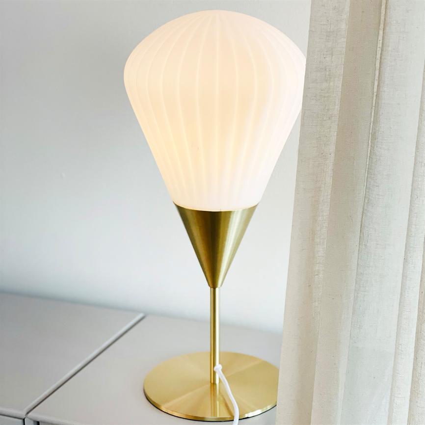 Bordlampe med opalhvid, ribbet skærm lyser blødt; skærmen sidder på et gyldent konisk stativ, lampen står på et lyst skab ved et gennemskinneligt hvidt gardin.