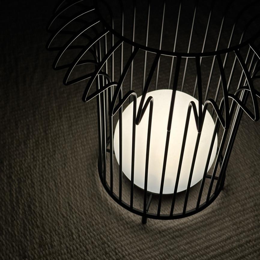 Spherical white lamp glows inside a decorative black metal cage, casting soft light on a bright, woven carpet in a dark room.