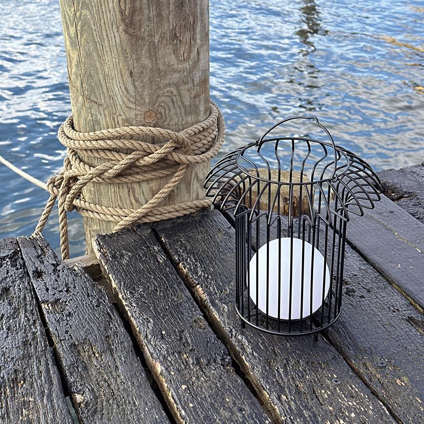 A black metal lantern with a white ball rests on a dark, worn wooden deck; close by stands a wooden pole wrapped in rope, with waves and water in the background.
