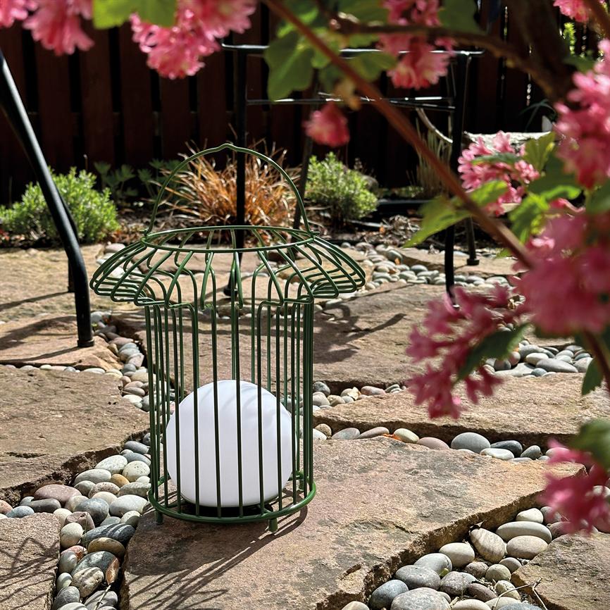 A green metal lantern shaped like a cage holds a white sphere, stands on flat stones between small round stones on a sunlit stone pavement, surrounded by pink flowers.