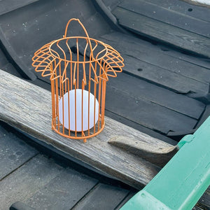 Orange wire cage lantern with white ball light source stands on a worn wooden board in a black rowing boat, surrounded by black seats and a green painted railing.