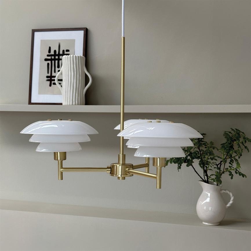 Brass ceiling lamp with three layered white shades hangs above a low shelf in front of a beige wall with framed black and white graphics, a white vase and a small plant.