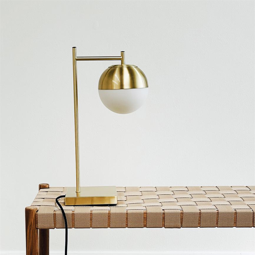 Gold colored table lamp with hemispherical opal glass shade stands on a woven wooden bench; arm holds the ball screen out from the base in a bright, minimalist space, with visible black cord.