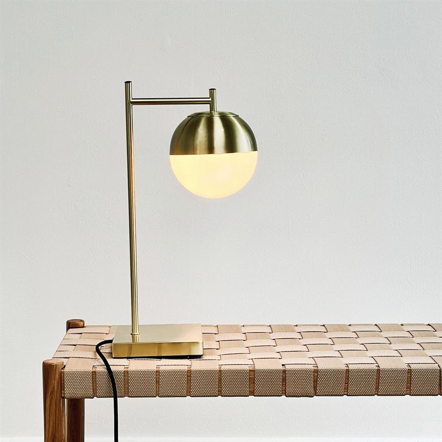 A brass table lamp with hemispherical lighted glass and metal shade sits on a vertical pole; it stands on a wicker light brown bench with black cord in front of a white wall.