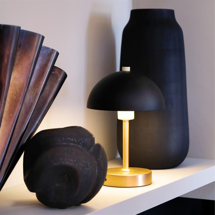 A small table lamp with a black dome and gold-coloured base emits warm, soft light; it stands on a shelf between a dark vase and a black sculpted stone in front of a light wall.