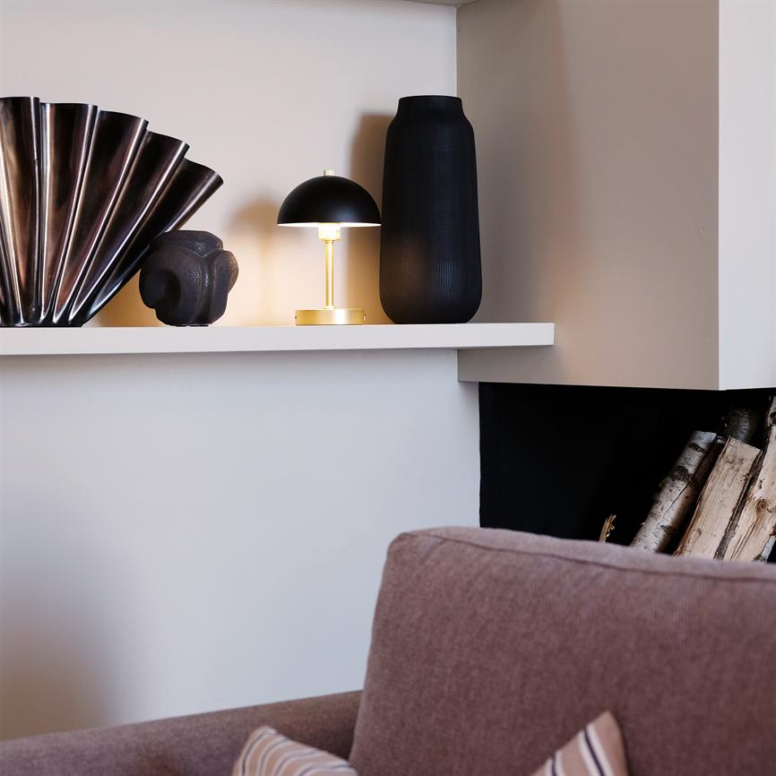 A small black and yellow table lamp shines softly on a white shelf between a folded metal vase, a dark sculpture and a tall black vase, above a pink sofa and stacked firewood.