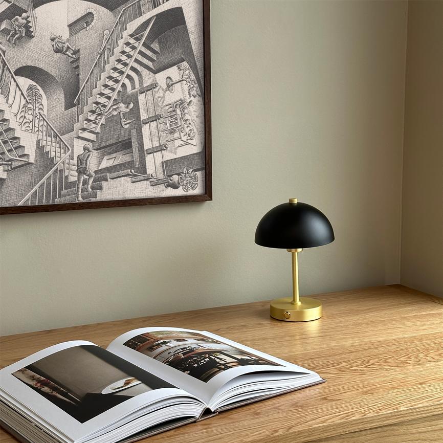 A black and gold table lamp is switched off next to an open photo book on a light wooden desk, under a framed Escher-like graphic poster on a beige wall.