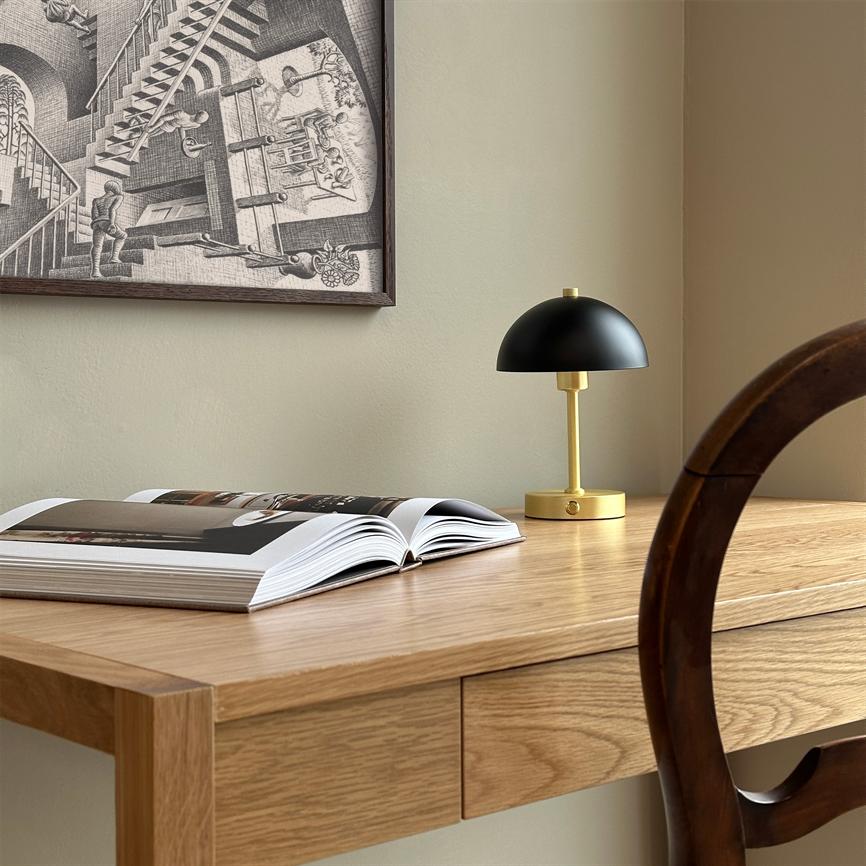 An open book lies on a light wooden desk; a small black-and-gold table lamp is switched off next to it, while a graphic black-and-white poster hangs on the wall and a wooden chair casts a shadow.