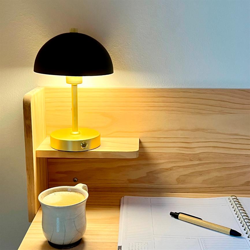 A yellow table lamp with a black dome shines warmly above a light wooden desk; a ceramic cup with coffee is on the left, and an open spiral notebook with a pen is on the right.