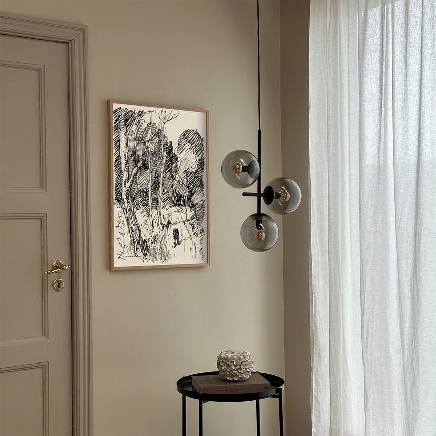 A framed black-and-white sketch of trees hangs on a beige wall; a three-globe glass pendant hangs next to it, and a small round side table with book and decorative object stands by a bright curtain.
 Text: KINFOLK