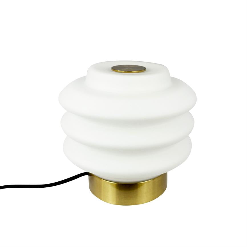 Bordlampe med hvid, bølget skærm og rund messingfod står på hvid baggrund; en sort ledning løber ud til venstre.