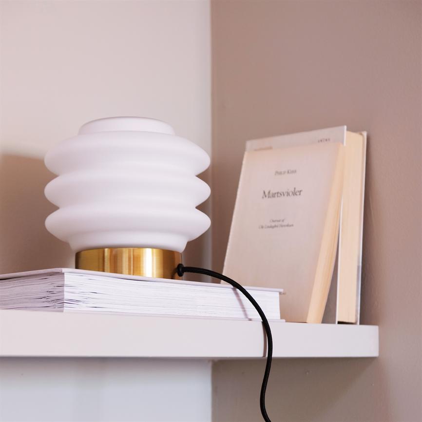 A white, wavy table lamp stands on a stack of books on a white shelf; a beige book stands upright behind against a light brown wall. 
 Text on the book cover: "March violets" (other text not clear).