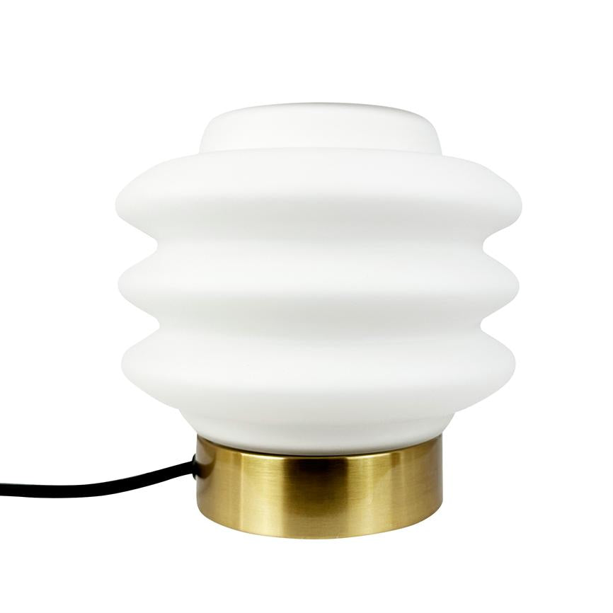 En hvid, bølget bordlampe hviler på en rund, børstet messingfod; sort strømkabel løber ud fra siden mod en neutral hvid baggrund.