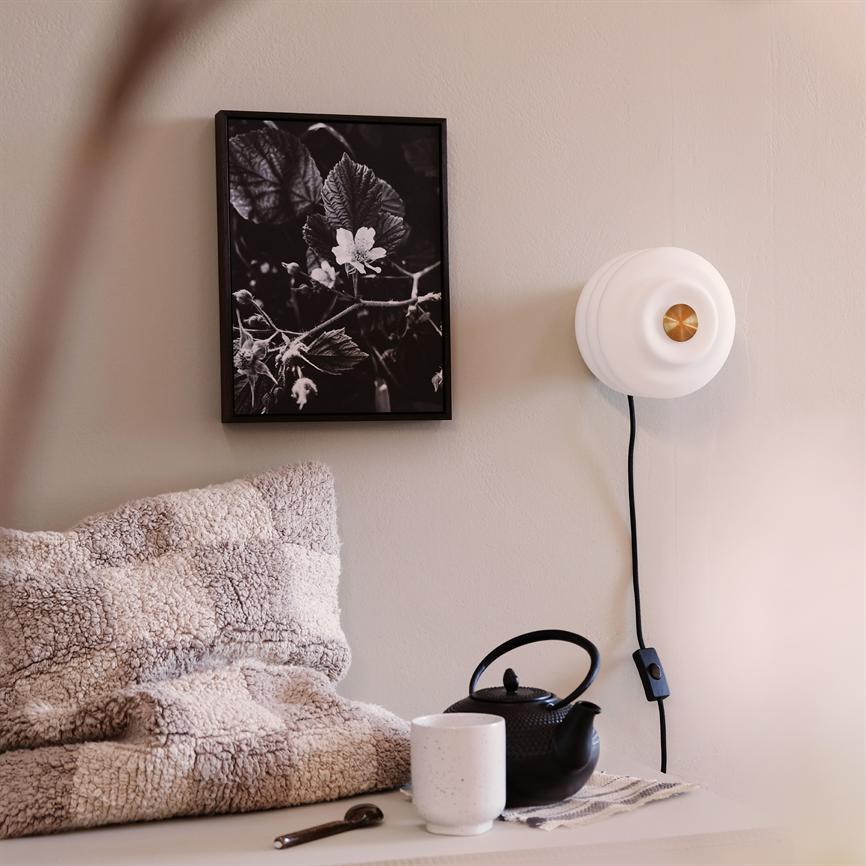 A framed black-and-white flower photograph hangs quietly on the wall, flanked by a white wall lamp; in front is a soft plaid, a black teapot and a white cup on a shelf.