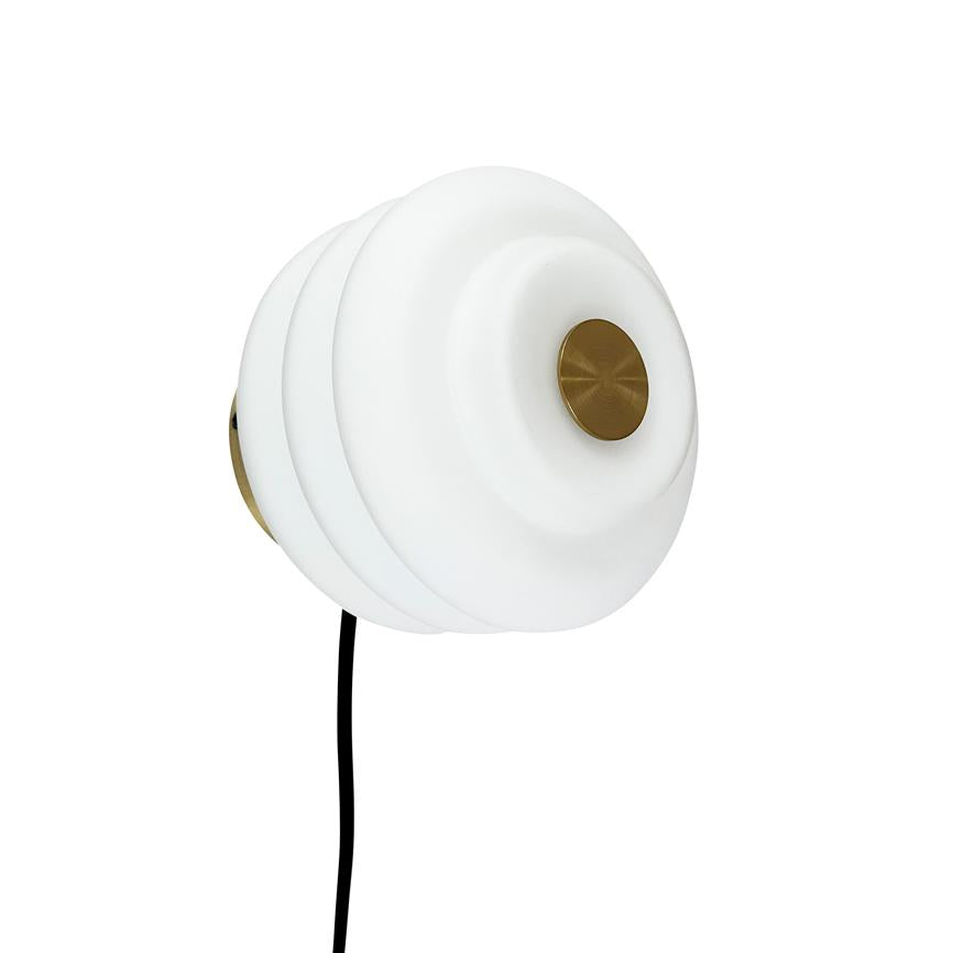 Wall lamp with round, layered white shade and central brass knob; hanging from a black cord in front of a uniform white background.