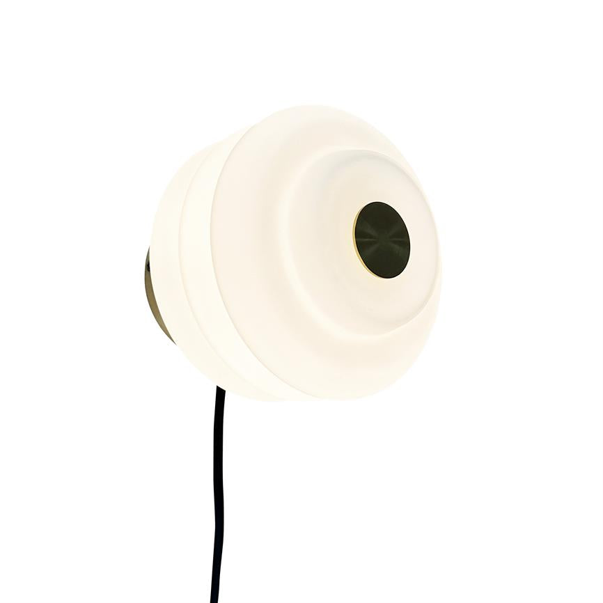 Round wall lamp glows dimly; layered white screen with a dark, metallic circle in the center. Black cord hangs down from the lamp, mounted against a neutral white background.