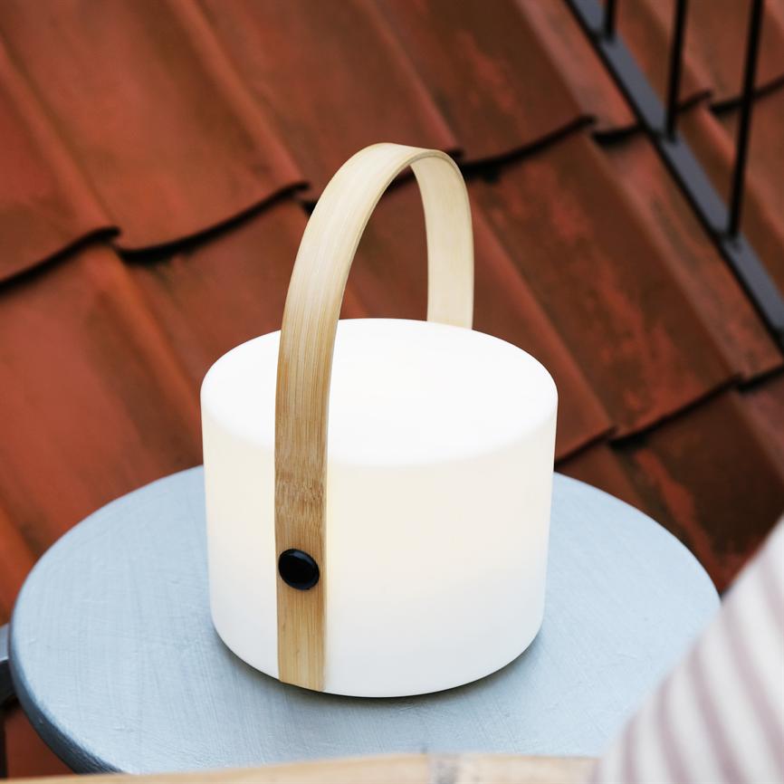 White cylindrical lamp with curved wooden handle stands on a small blue round table. It emits soft light, placed in front of red roof tiles in an outdoor balcony environment.